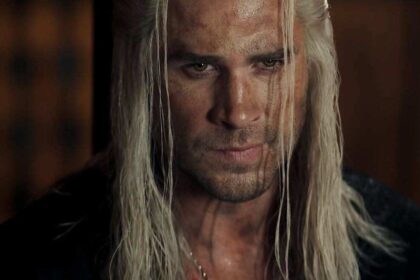 लियाम हेम्सवर्थ ‘The Witcher’ में Geralt के रूप में तलवार के साथ खड़े दिख रहे हैं, डार्क जंगल के बैकग्राउंड में।